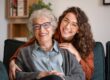 Smiling elderly woman and younger woman sitting close together on a couch, both wearing glasses and looking happy while embracing each other warmly.