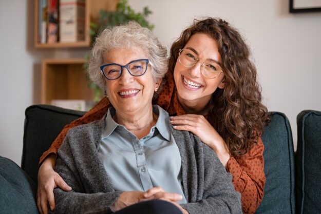 Smiling elderly woman and younger woman sitting close together on a couch, both wearing glasses and looking happy while embracing each other warmly.