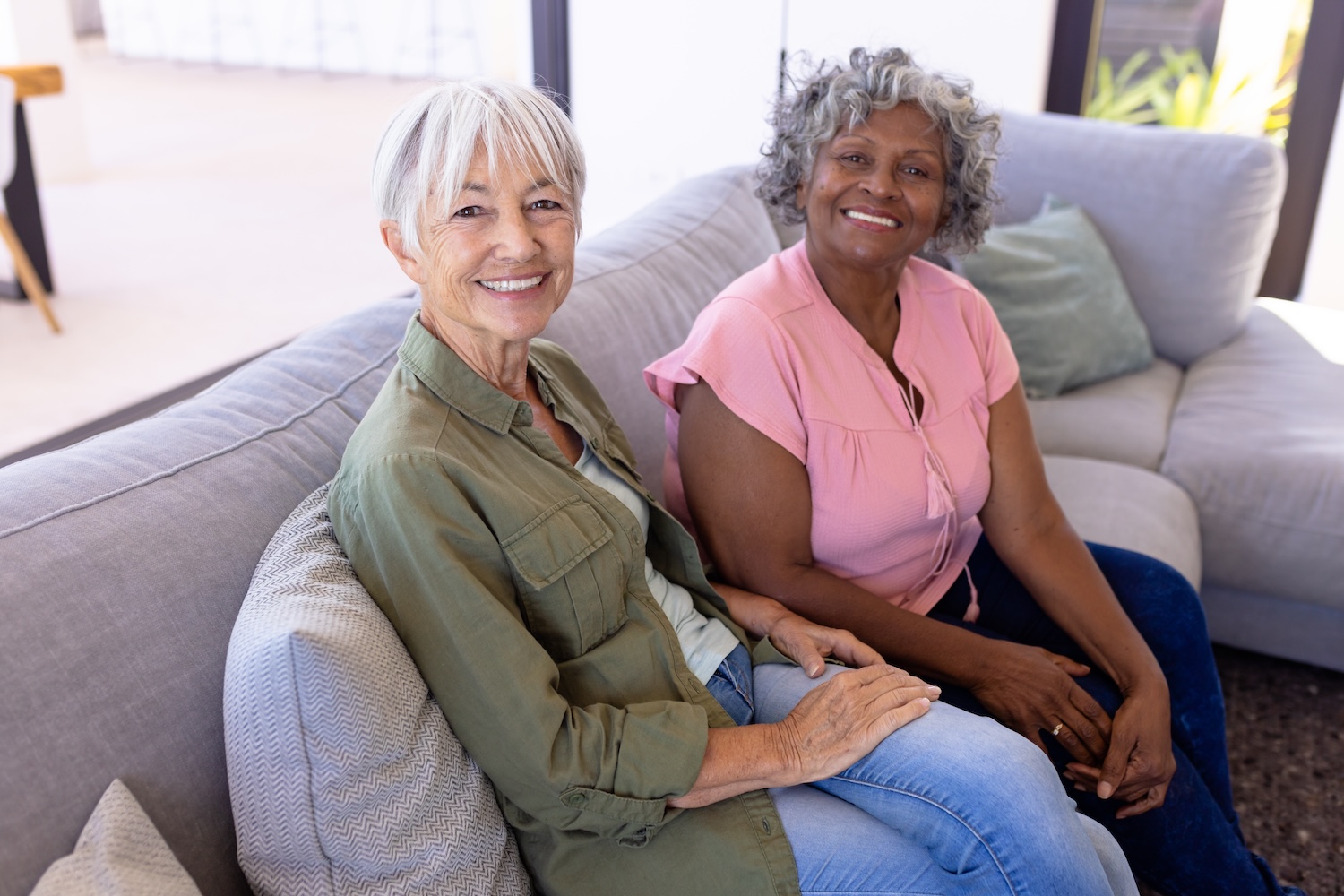 Your Guide to Respite Care in Rochester MI Two older women sitting close together on a couch, smiling warmly at the camera in a bright living room.